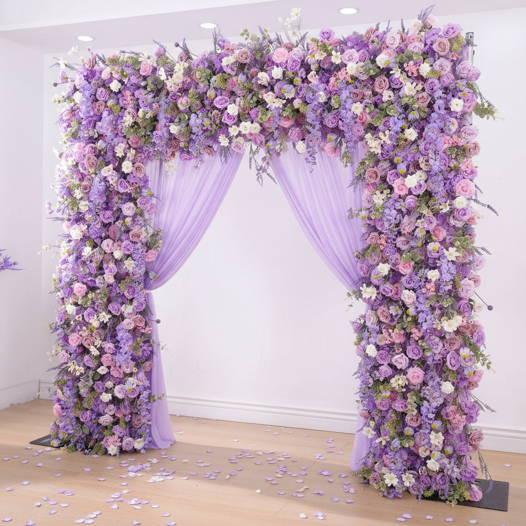 Woman in a white dress standing under a floral arch decorated with purple and pink flowers.